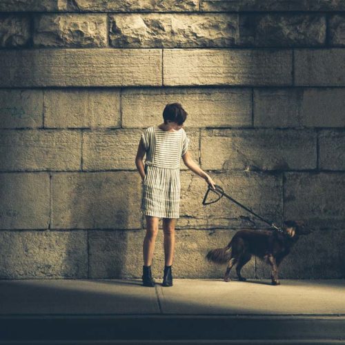 A woman standing in a striped jumper suit holding a leash with a dog walking to the right of the picture. The concrete alleyway has a shadow on it with a rock wall in the background. The dog owner is thinking about the downtown Austin, TX dentists office at Toothbar.