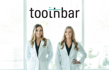 An image of both doctors standing in doctor jackets in front of a floor to ceiling window in the Toothbar downtown Austin dentistry. Both doctors are smiling and ready to take on new dental patients needing Invisalign®, cosmetic dentistry like veneers or crowns. As well as offing other services, like the amazing Toothbar lounge.