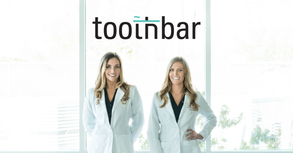 An image of both doctors standing in doctor jackets in front of a floor to ceiling window in the Toothbar downtown Austin dentistry. Both doctors are smiling and ready to take on new dental patients needing Invisalign®, cosmetic dentistry like veneers or crowns. As well as offing other services, like the amazing Toothbar lounge.