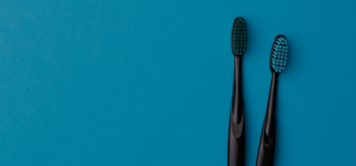 A blue background with two black toothbrushes on the right side of the image where each toothbrush has colored bristles. The toothbrush on the left has green bristles and the toothbrush on the right has blue bristles. Each toothbrush has a black casing.